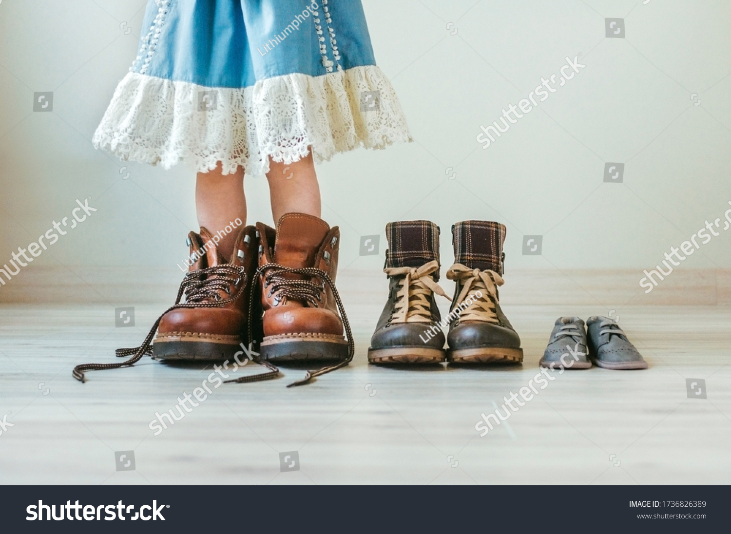 Close up of little girl putting on fathers hiking shoes. Three pairs of shoes for family indoor. #1736826389