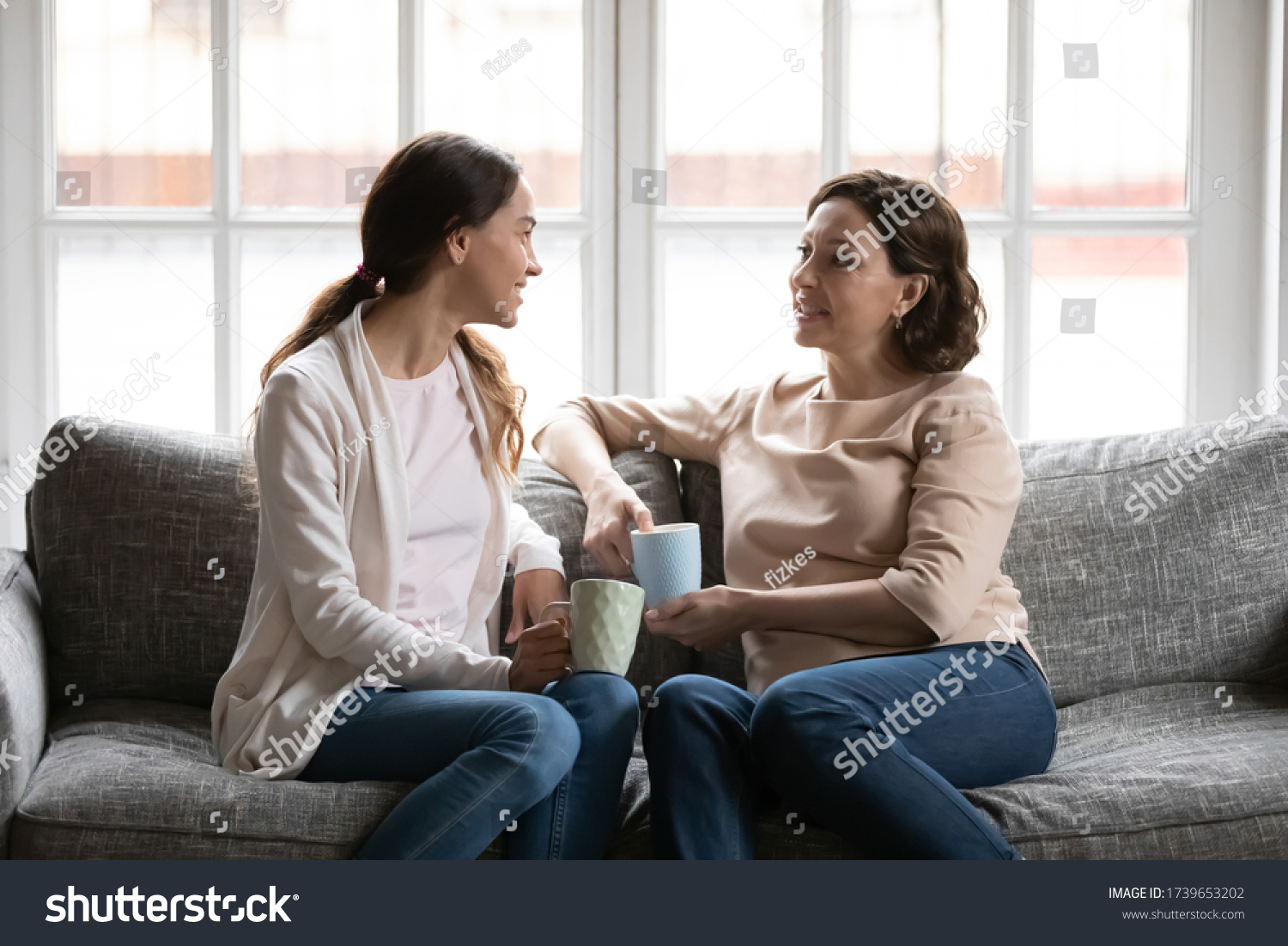 Happy middle-aged mother and adult daughter sit on couch in living room talk drinking tea or coffee, smiling senior mom and grownup millennial girl have fun relax on sofa ta home chatting gossiping #1739653202