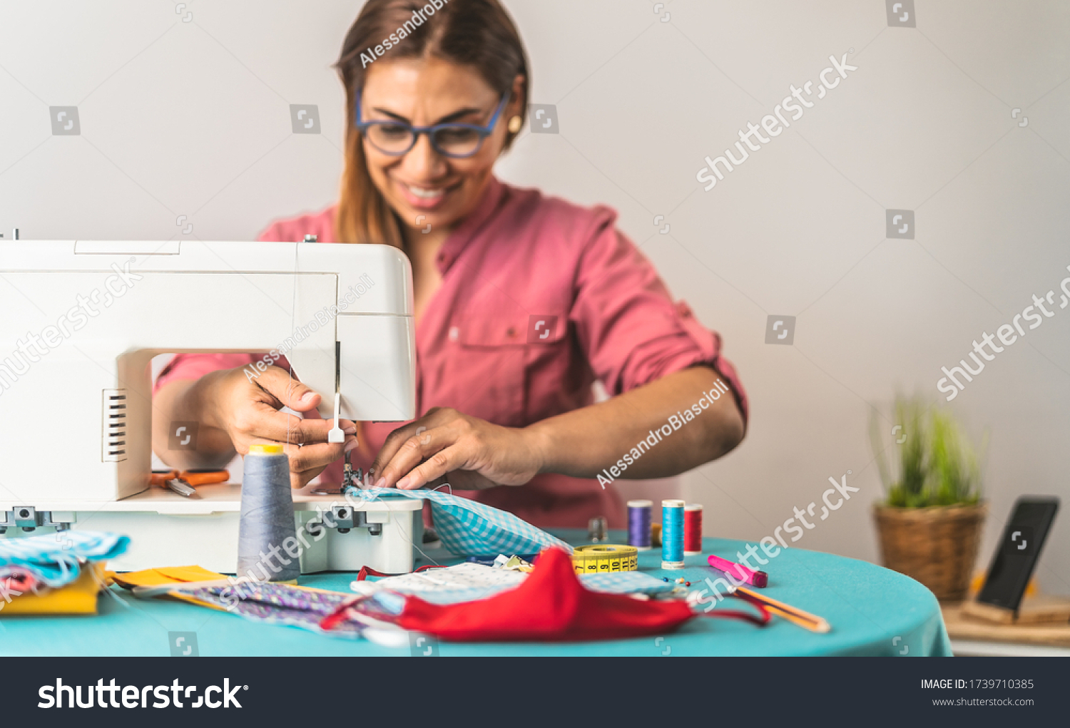 Happy Latin tailor seamstress woman sewing with machine homemade medical face mask for preventing and stop corona virus spreading - textile industry and covid19 healthcare concept  #1739710385