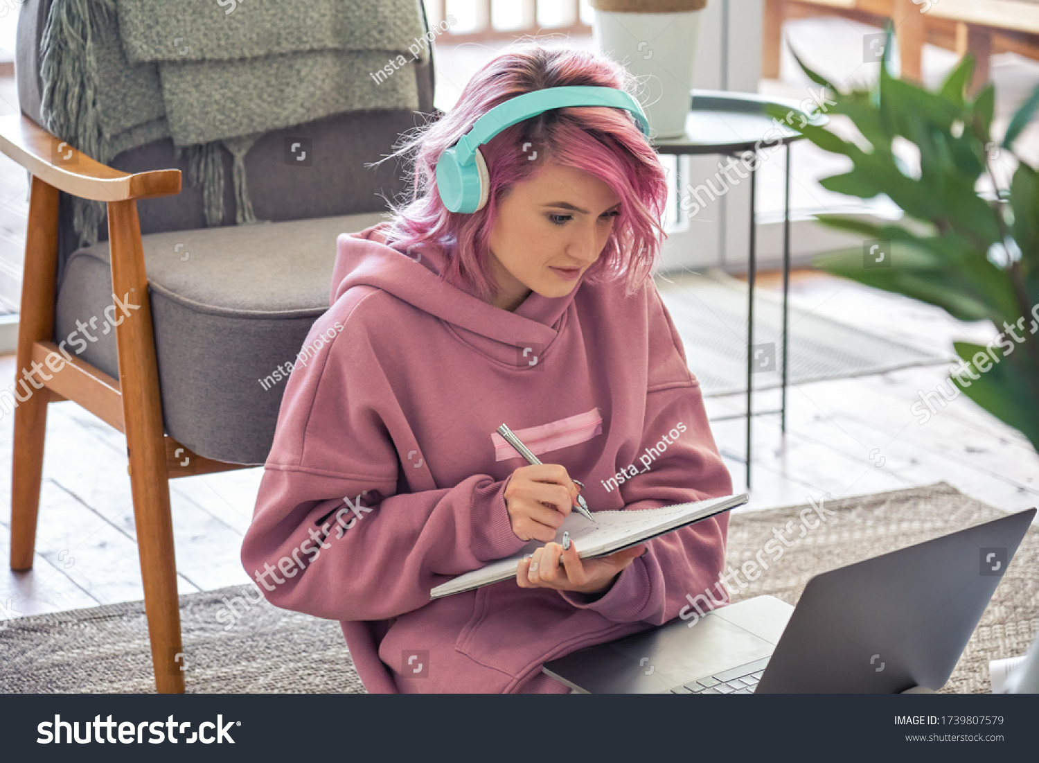 Focused hipster teen girl school college student pink hair wear headphones write notes watching webinar online video conference calling on laptop computer sit on floor working learning online at home. #1739807579