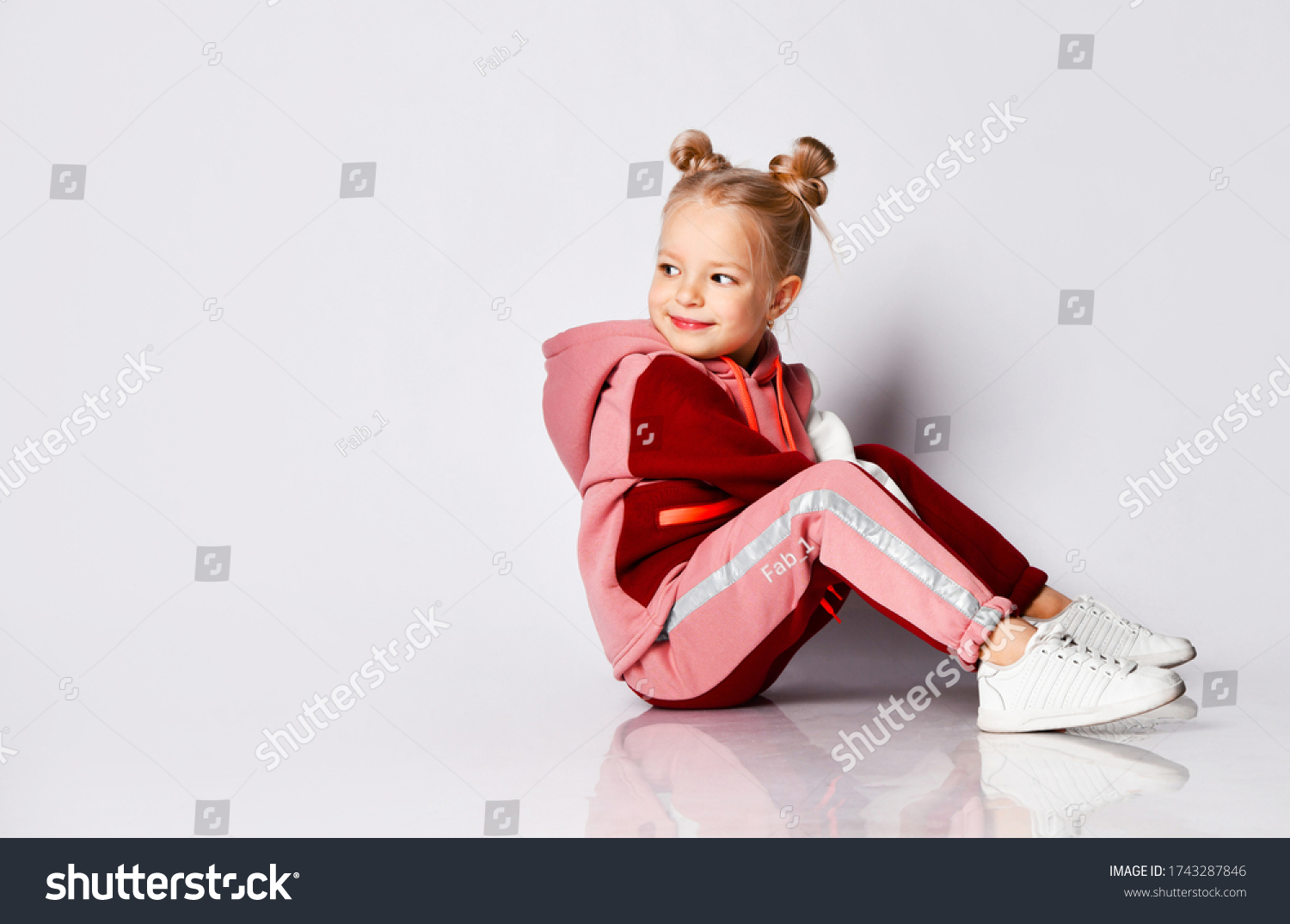 Little blonde girl with buns hairstyle, in colorful tracksuit, sneakers. She laughing, sitting on floor, posing isolated on white. Childhood, fashion, advertising and sport. Close up, copy space #1743287846