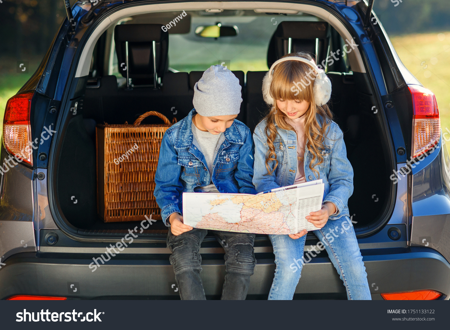 Cute stylish boy and pretty 10-s girl looking at the road map while sitting in the auto's trunk and discussing right direction to their family trip. Family vacation trip by car. #1751133122