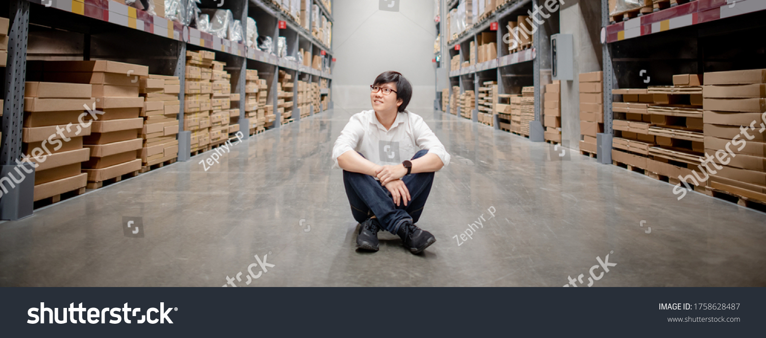 Asian shopper man sitting between cardboard box shelves aisle in warehouse choosing what to buy. Shopping lifestyle in department store. Buying or purchasing factory goods. Inventory industry concept #1758628487