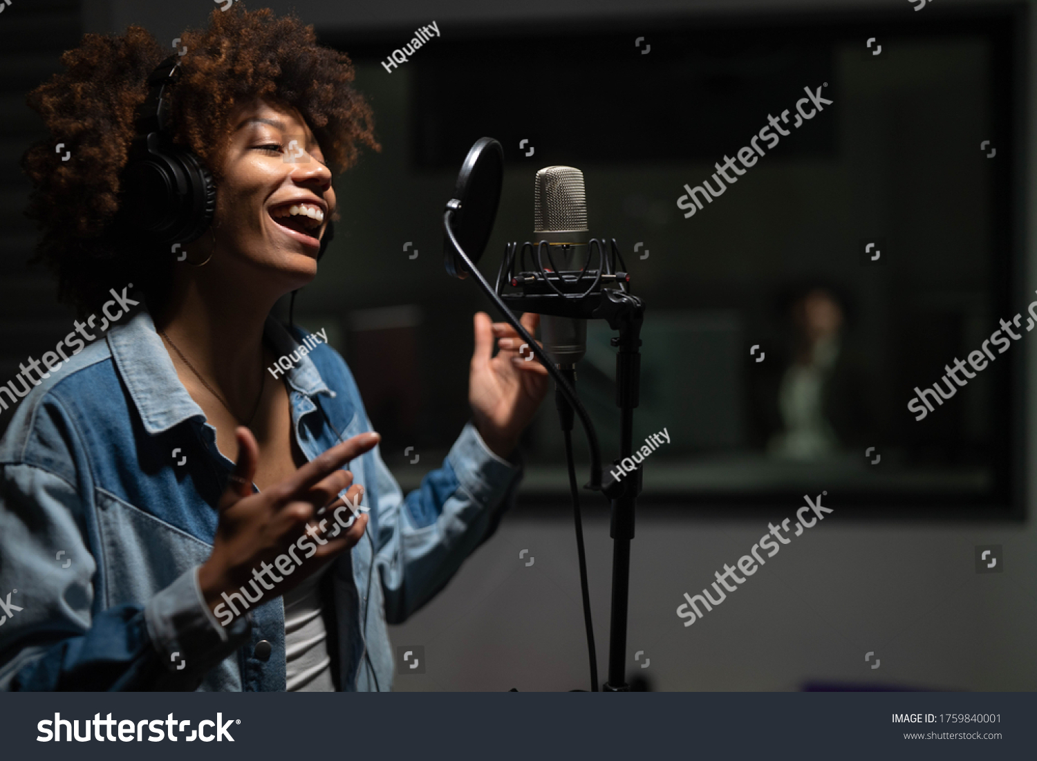 An young professional smiling energetic african female singer wearing headphones is performing a new song with a microphone while recording it in a music studio.  #1759840001