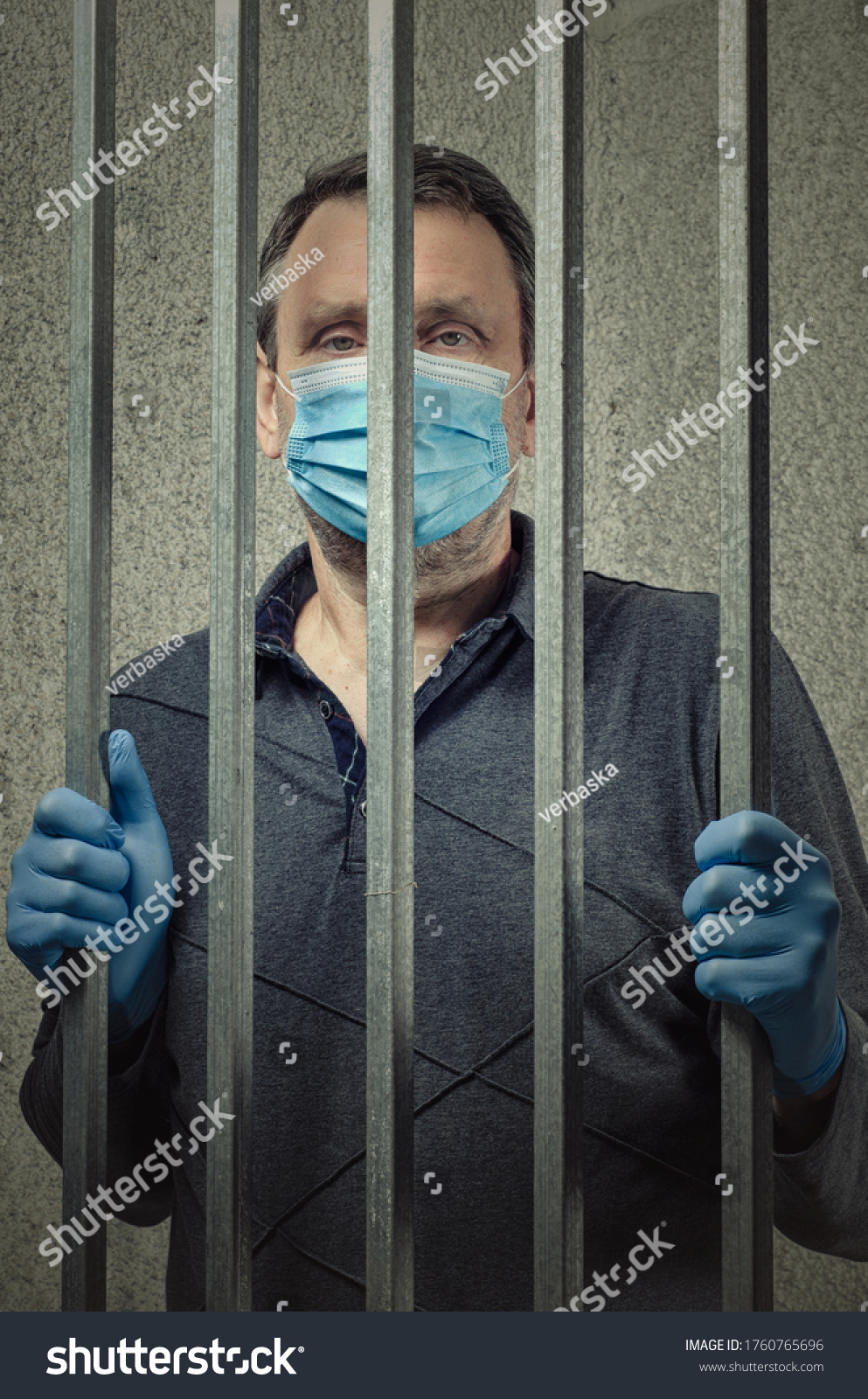 A middle-aged man in a blue medical face mask and blue gloves is looking through the bars of a pre-detention cell. He was arrested for a quarantine violation. #1760765696