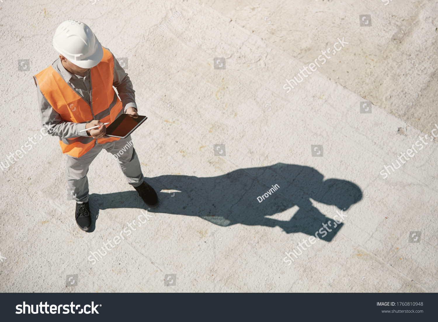An engineer in an orange vest and a white construction control helmet conducts an inspection with a tablet in his hands, top view #1760810948