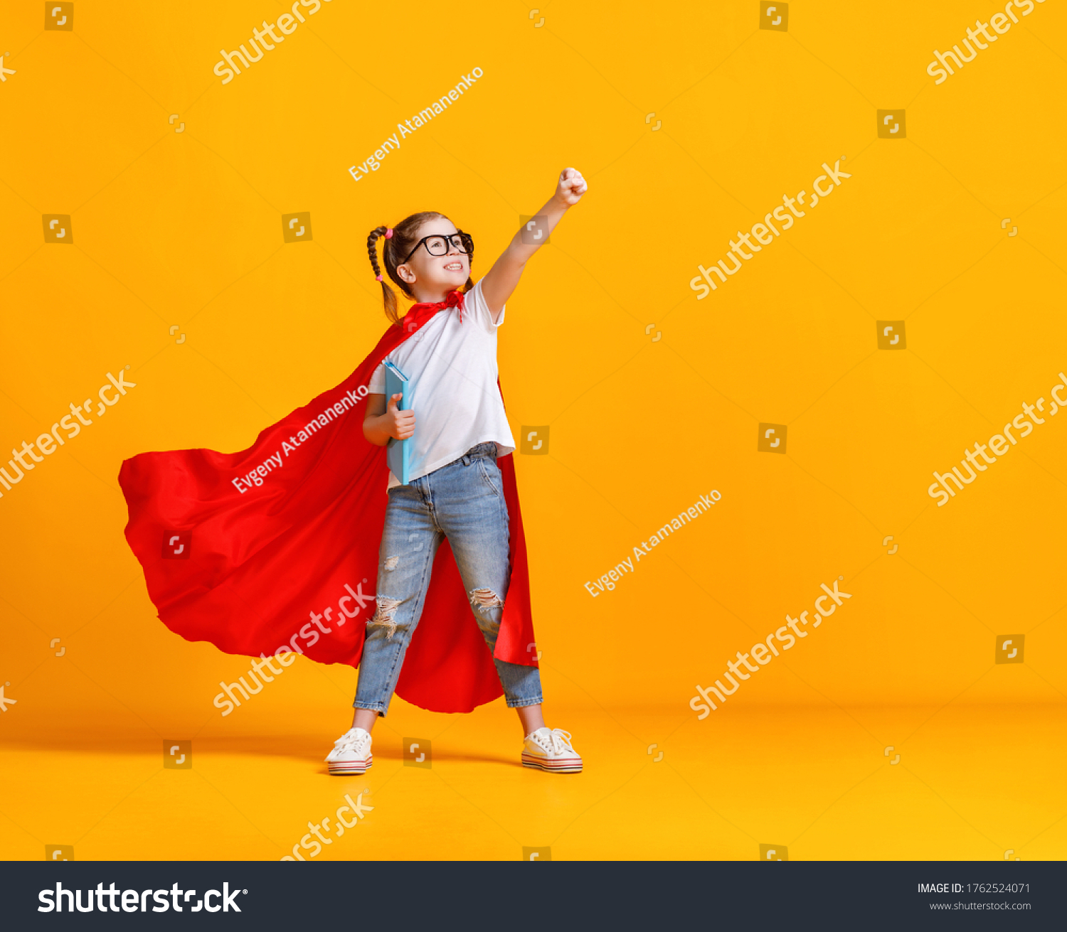 Full body girl in superhero cape smiling and raising fist up while being ready for school studies against yellow backdrop
 #1762524071