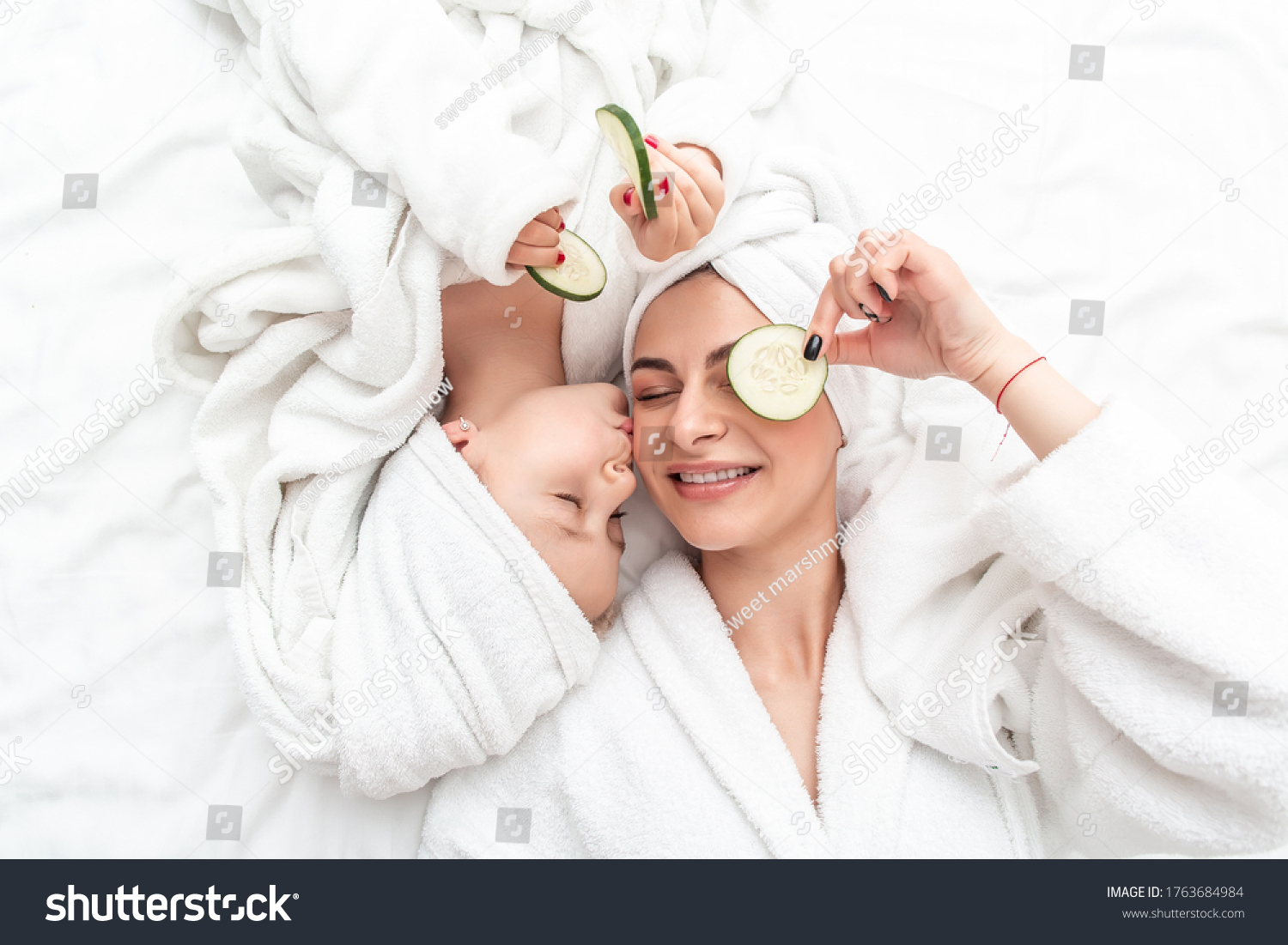 Mom and daughter in the bedroom doing face mask applying pieces of cucumber to their eyes. Mother with child doing beauty treatment together. #1763684984