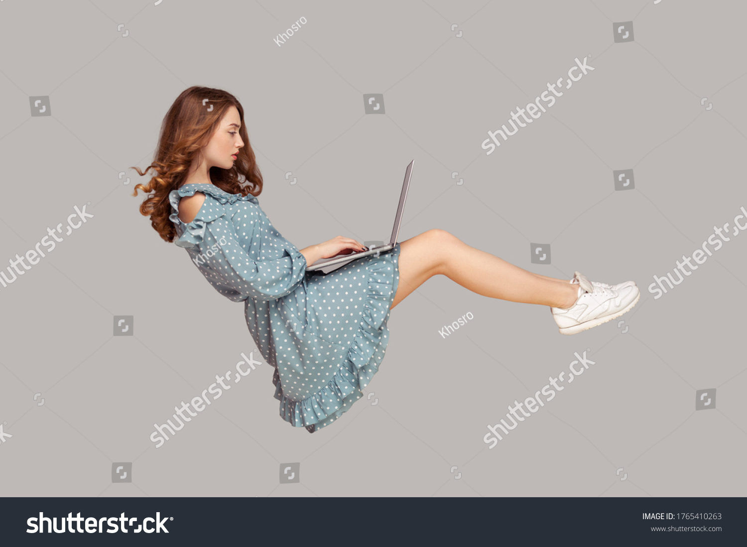 Hovering in air. Pretty girl ruffle dress levitating, typing keyboard using laptop for work online, surfing web social networks while flying in mid-air. indoor studio shot isolated on gray background #1765410263