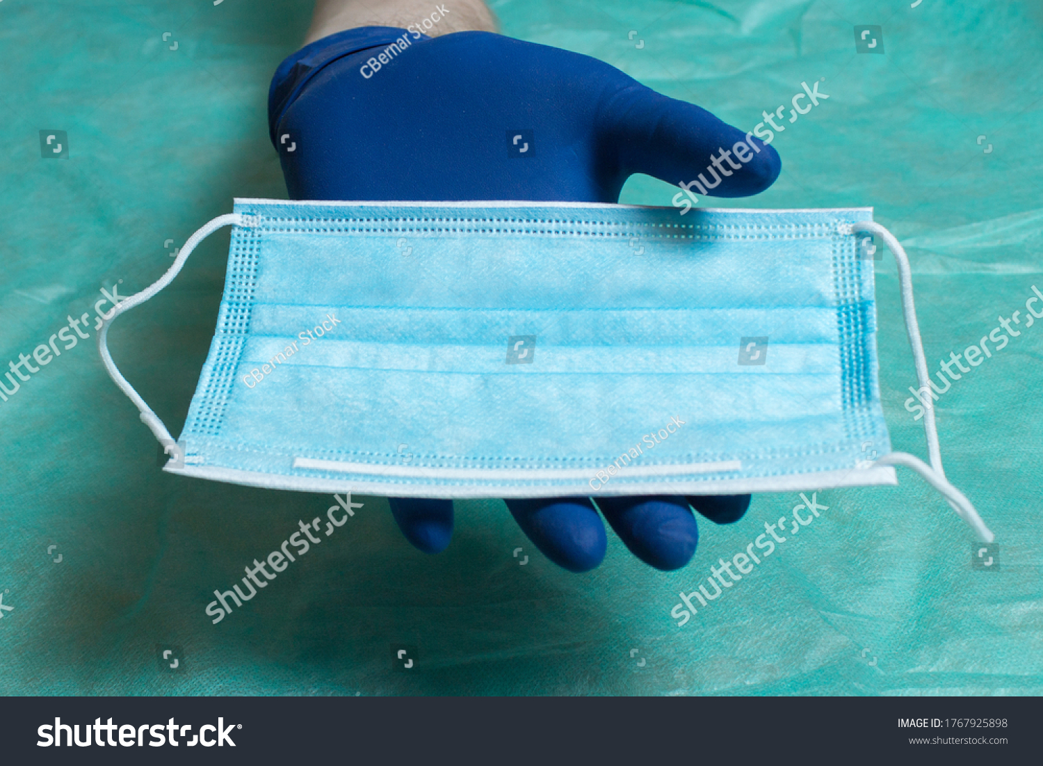 A hand protected with blue gloves shows a facial mask in the palm of his hand on a green cloth of medical surgical material. Health care and surgical concept. Personal protective
equipment. #1767925898