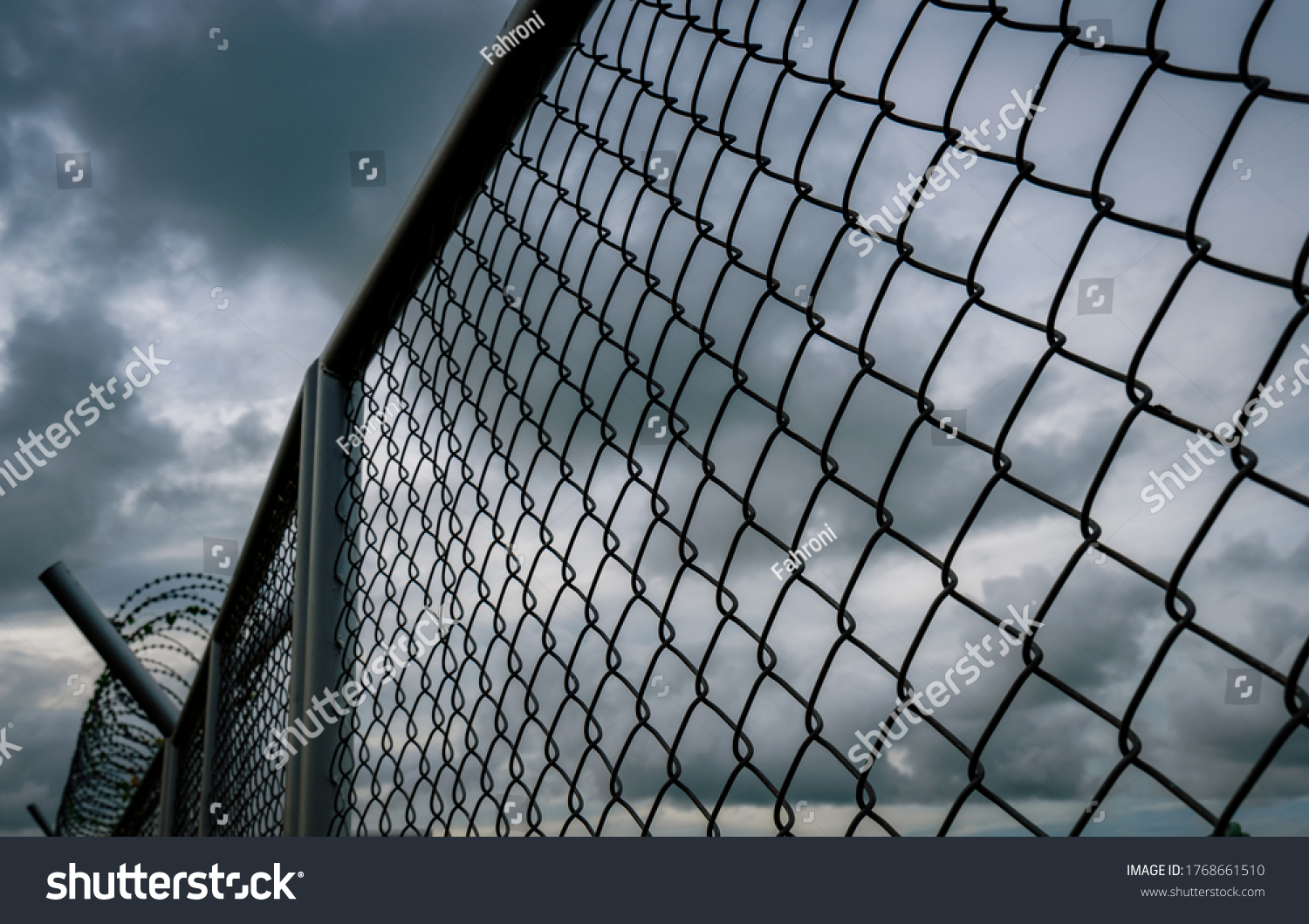 Military zone mesh fence. Prison security fence. Looking up view of barbed wire security fence with storm sky and dark clouds. Razor wire jail fence. Barrier border. Boundary security wall.  #1768661510