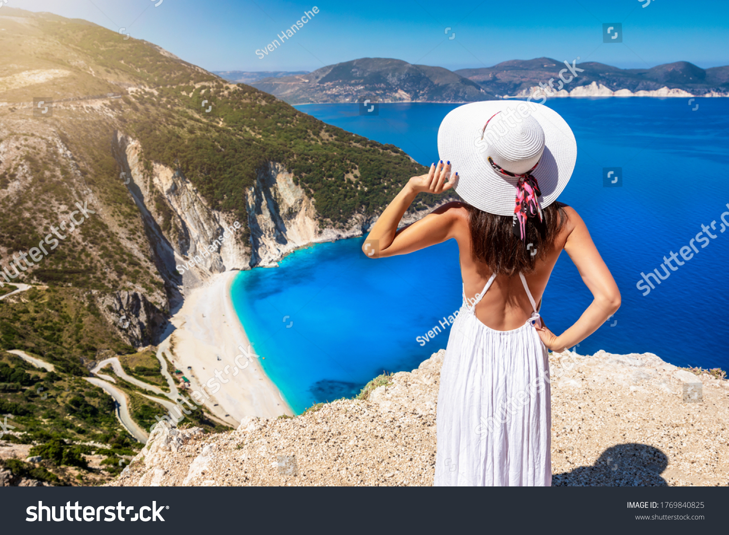 A beautiful tourist woman in white dress and sunhat enjoys the view to the famous Myrtos Beach on the island of Kefalonia, Ionian Sea, Greece #1769840825