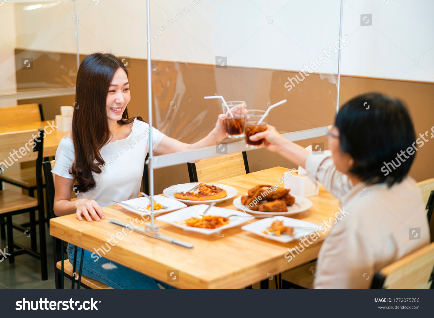 Asian beautiful female clinking a glass of water with her mother who siting separate and keep distance with table plastic shield partition in restaurant, new normal and social distancing concept #1772075786