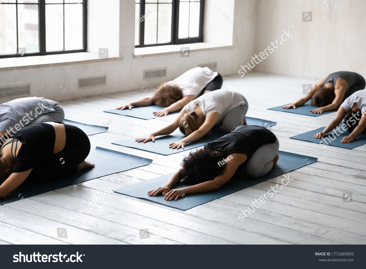 Side view multiracial young fit diverse people lying on floor mat in balasana position, enjoying restorative child pose together at group morning class in modern yoga club interior, relieving muscles. #1772685005