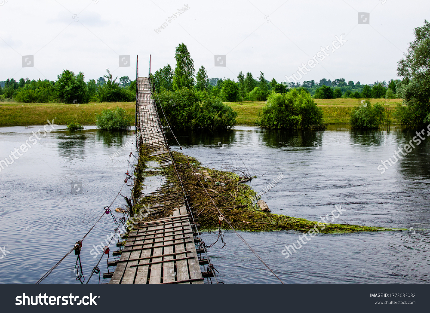 Suspension bridge over the river is broken and flooded against the backdrop of a summer landscape #1773033032