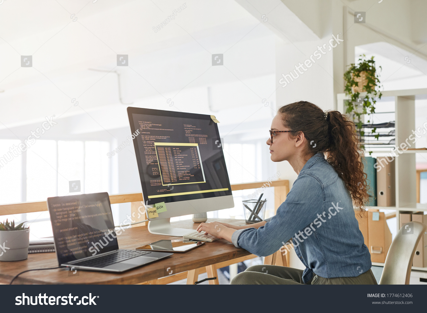 Side view portrait of female IT developer typing on keyboard with black and orange programming code on computer screen and laptop in contemporary office interior, copy space #1774612406