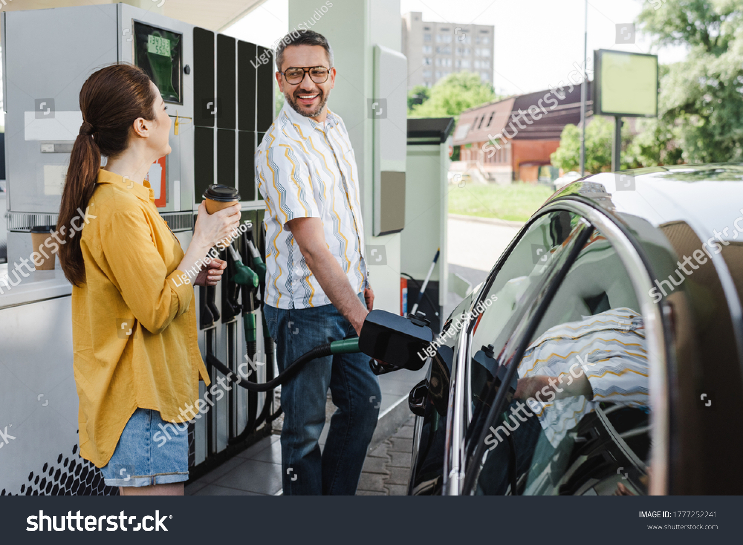 Selective focus of smiling man looking at wife with coffee to go while refueling car on gas station #1777252241