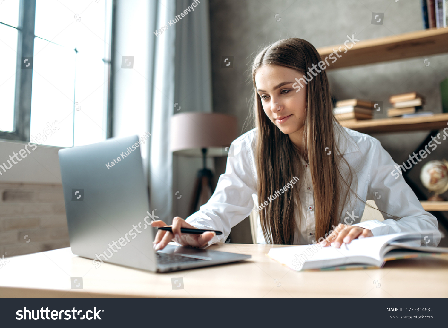A beautiful caucasian female student is studying in college remotely. She is sitting at a table at home with a laptop and a notepad and concentrated is watching a video conference lesson #1777314632