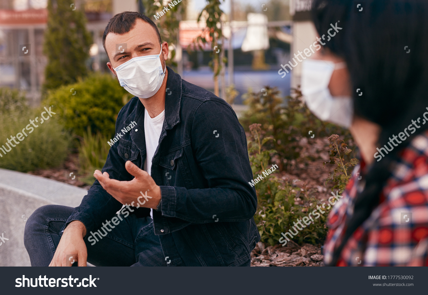 Young guy in medical mask gesticulating and talking with woman while sitting on border and keeping social distance during date on city street #1777530092