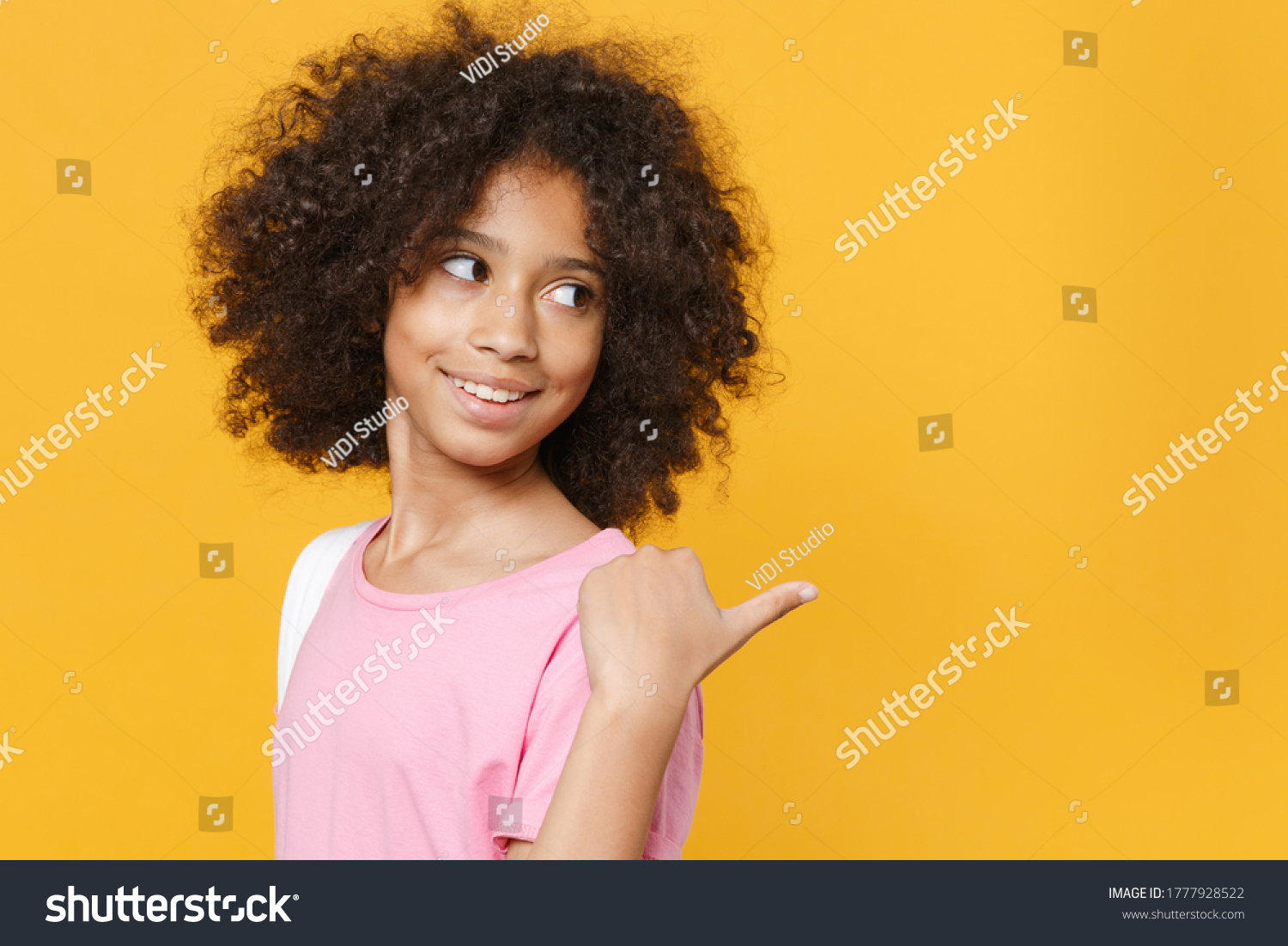 Side view of smiling little african american kid girl 12-13 years old in pink t-shirt isolated on yellow wall background studio. Childhood lifestyleconcept. Mock up copy space. Pointing thumb aside #1777928522