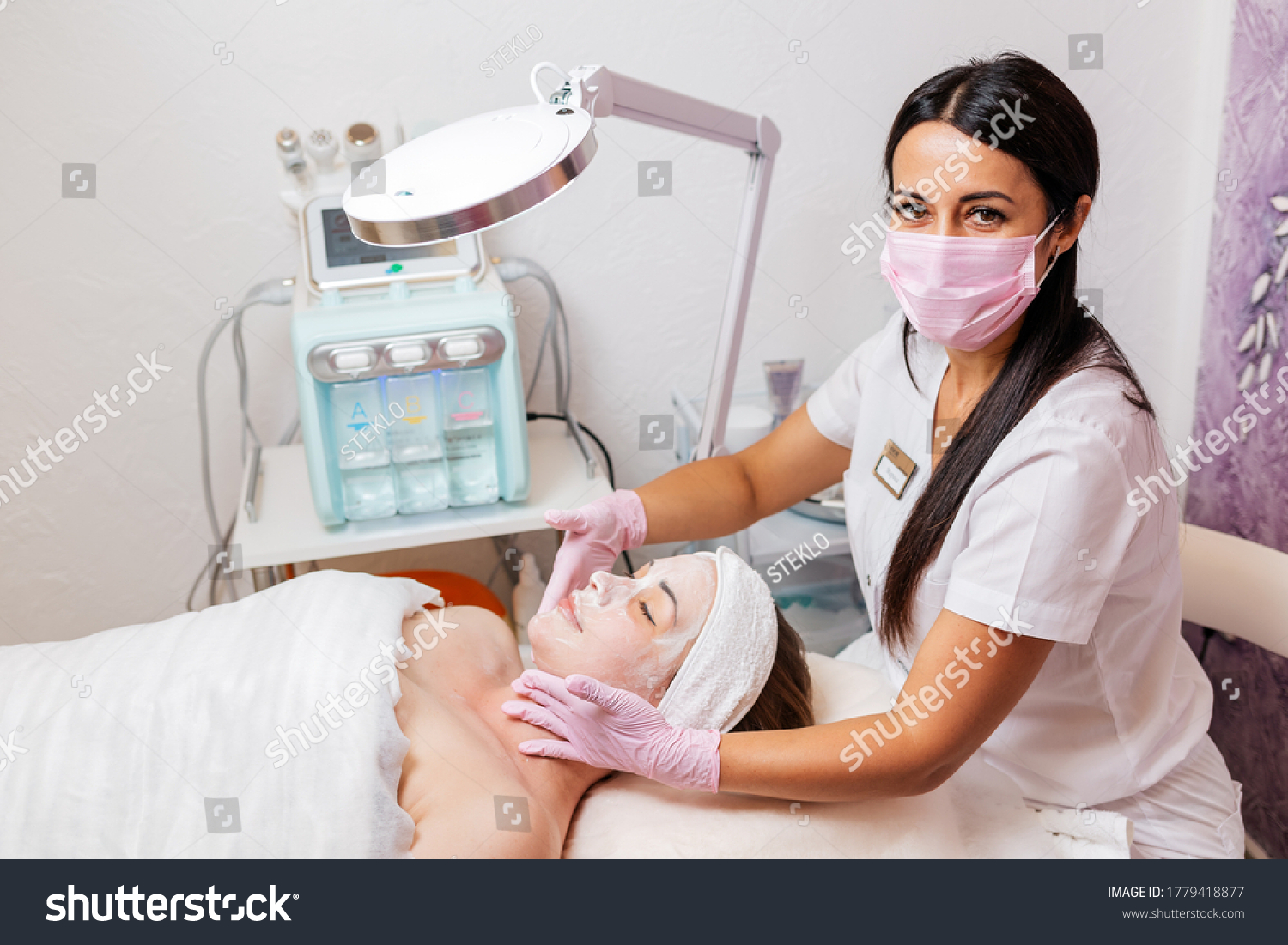 A cosmetologist in a medical mask and rubber gloves applies a cream mask to the client's face. Top view. In the background, a cosmetology device. Concept of cosmetology during the pandemic #1779418877