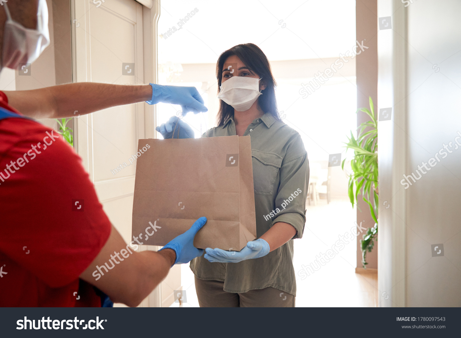 Indian woman customer wearing face mask and gloves taking delivery paper eco bag from man courier holding grocery food package delivering supermarket takeaway order standing at home. Safe delivery. #1780097543