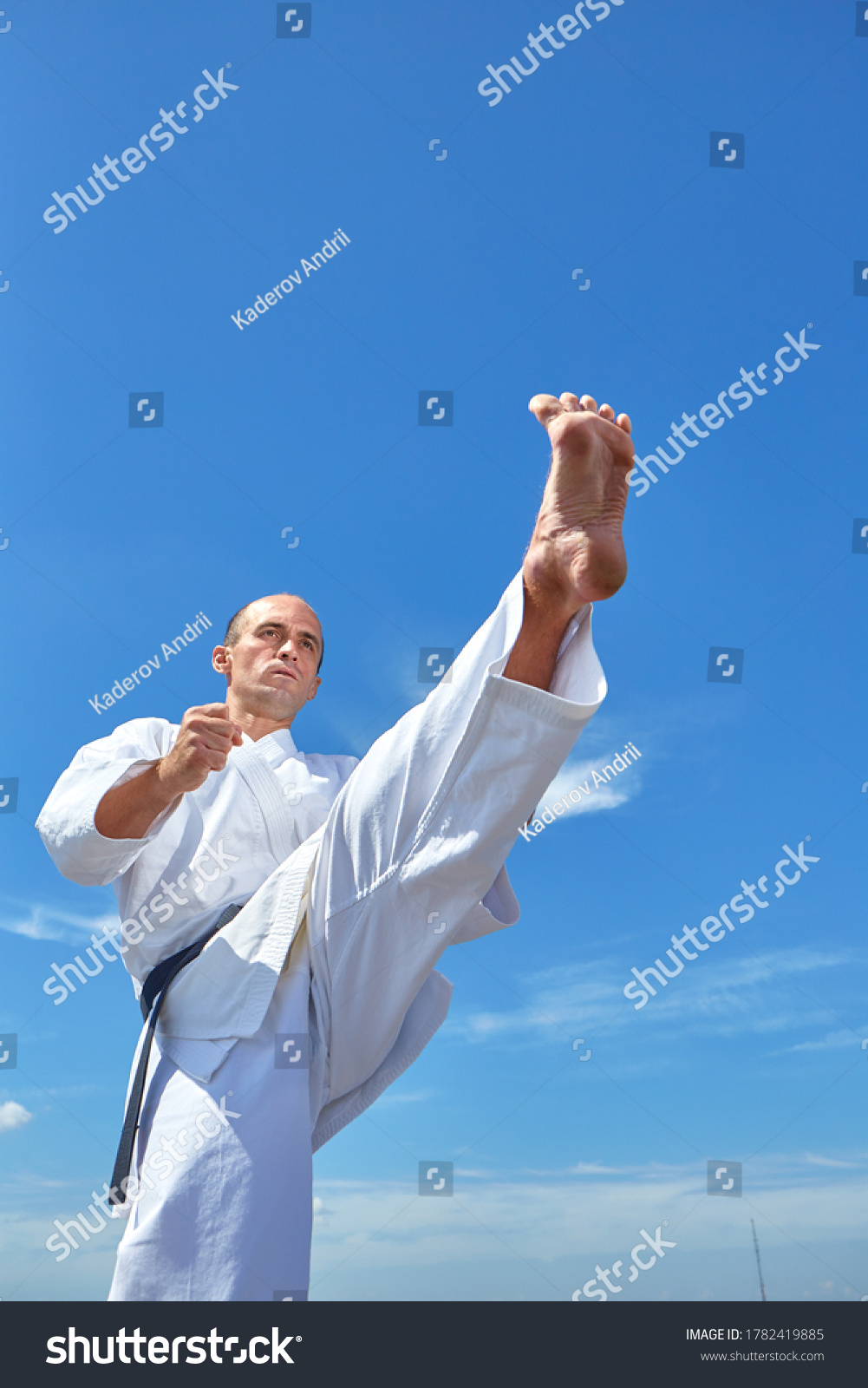 A sportsman in karategi trains a kick against a blue sky with clouds #1782419885