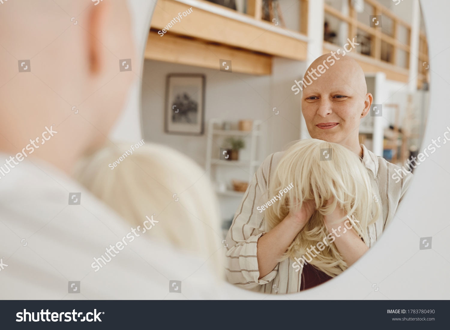 Reflection portrait of bald adult woman looking in mirror holding wig while standing in warm-toned home interior, alopecia and cancer awareness, copy space #1783780490