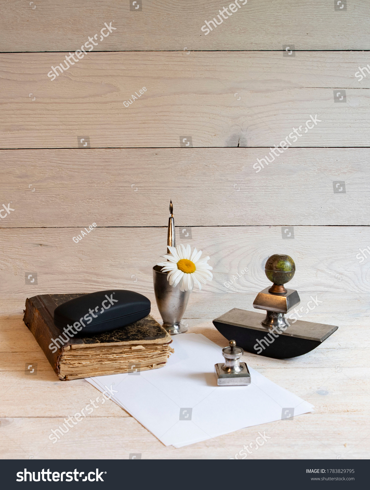 Old book, silver writing set of ink pens, paperweight, stamp and glass on a light table with white sheet of paper and chamomile #1783829795