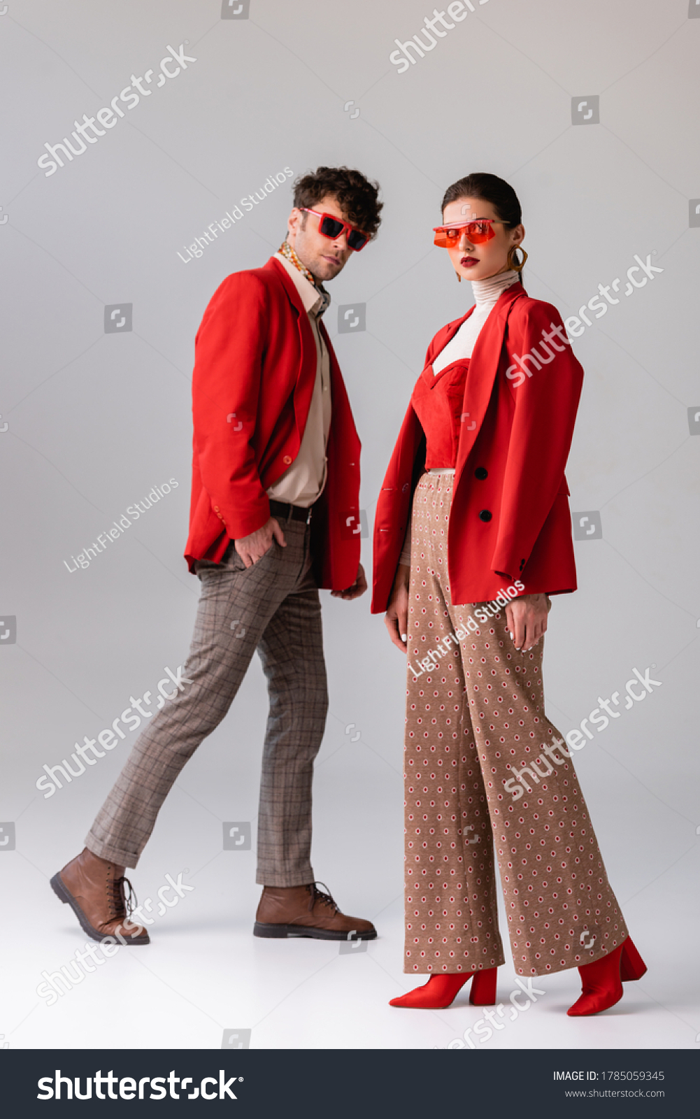 full length view of fashionable couple in red blazers and sunglasses looking at camera  on grey #1785059345