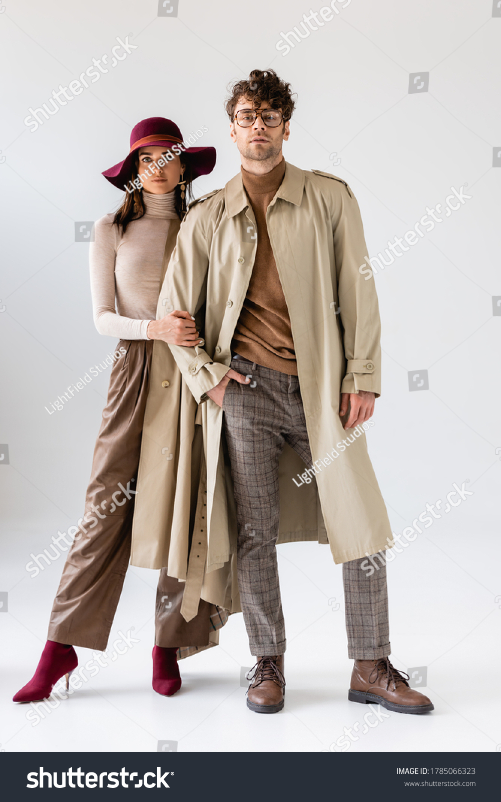 full length view of stylish woman in hat touching hand of trendy man in trench coat on grey #1785066323