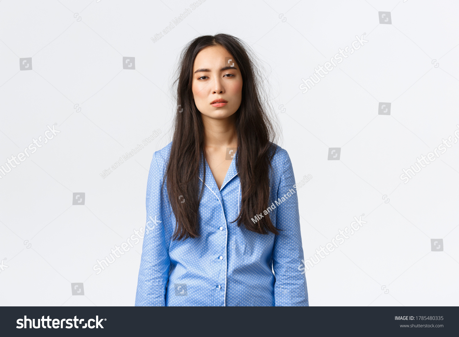 Exhausted asian woman with messy hair after lying in bed, wearing pajamas, looking tired with sleepy eyes as suffering insomnia, didnt have much sleep, waking up early, standing white background #1785480335