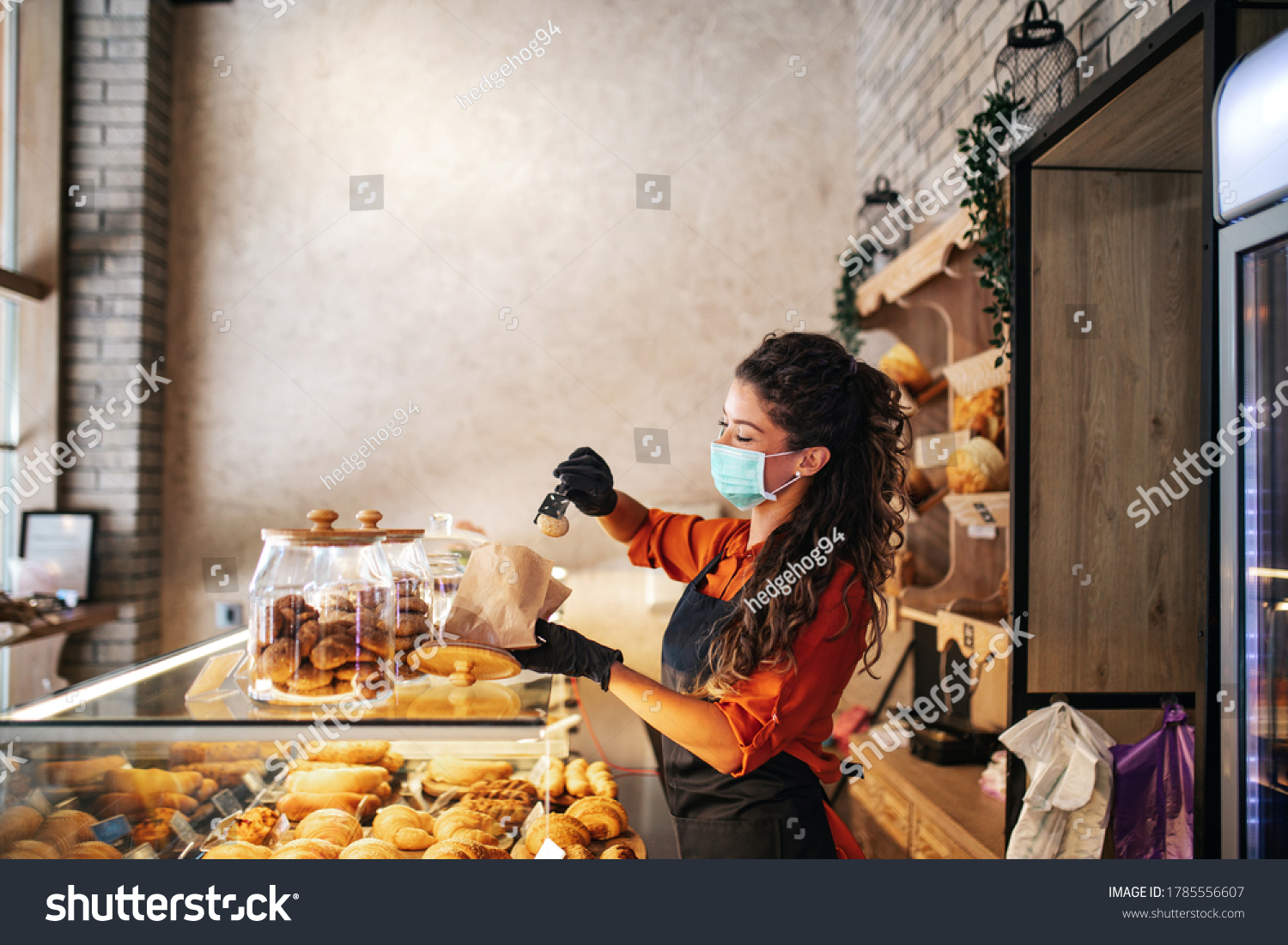 Beautiful young female worker with protective mask on face working in bakery. #1785556607