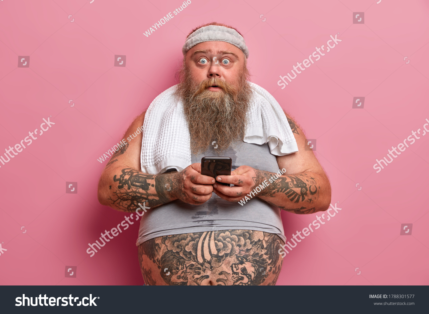 Chubby shocked man with thick beard, stares at camera, busy with sport training, dressed in sportswear cares about his weight uses cellular for checking how much calories were burnt. Sport, motivation #1788301577