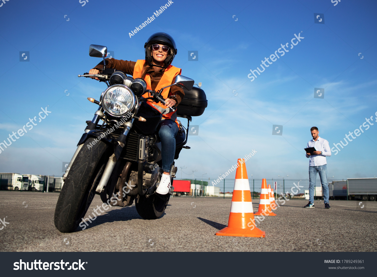 Motorcycle school of driving. Female driver with helmet taking motorcycle lessons and practicing ride. In background traffic cones and instructor with checklist rating and evaluating the ride.  #1789249361