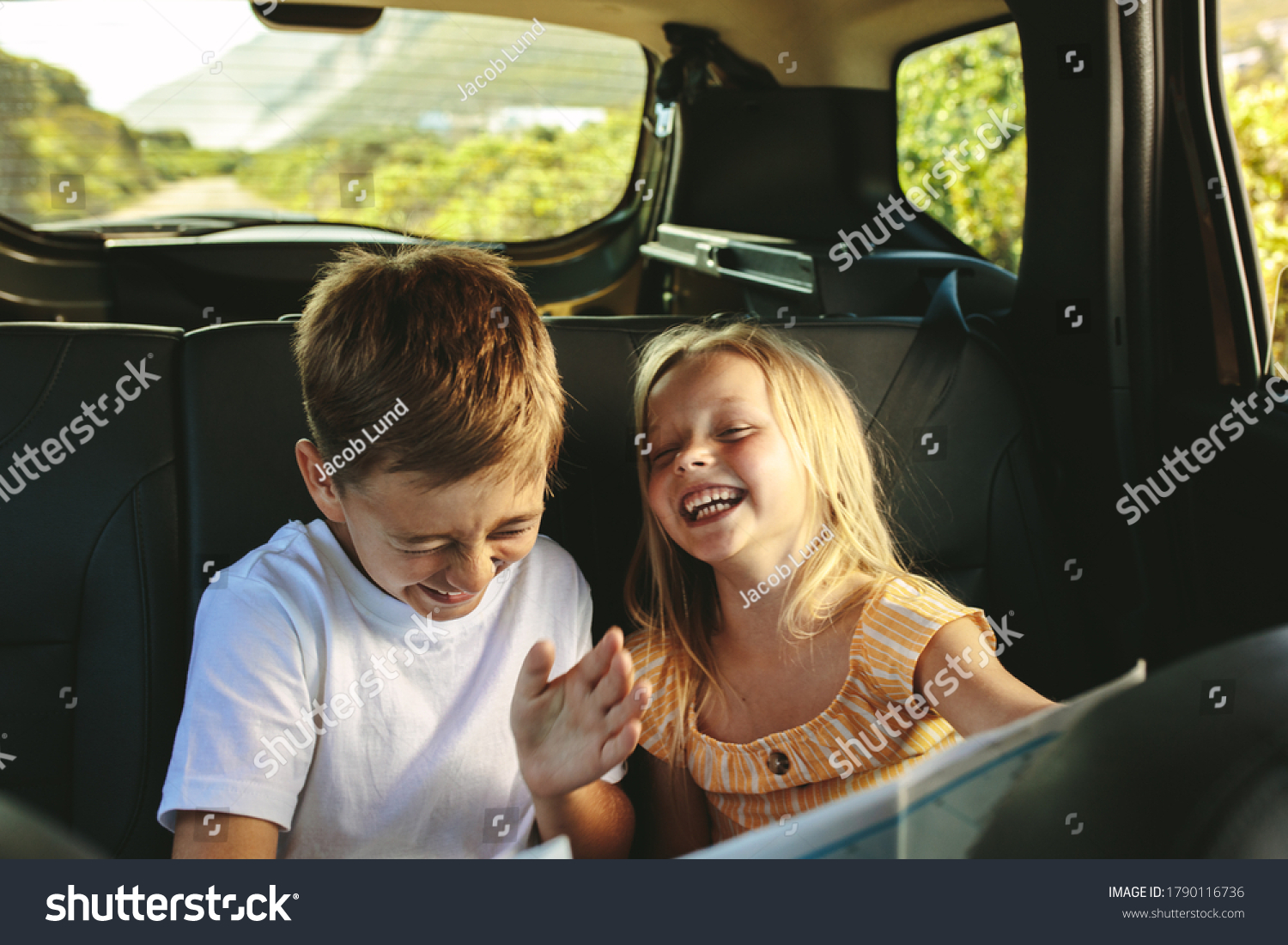 Siblings sitting on backseat of car looking at map and smiling. Kids traveling in a car on roadtrip playing with a map. #1790116736