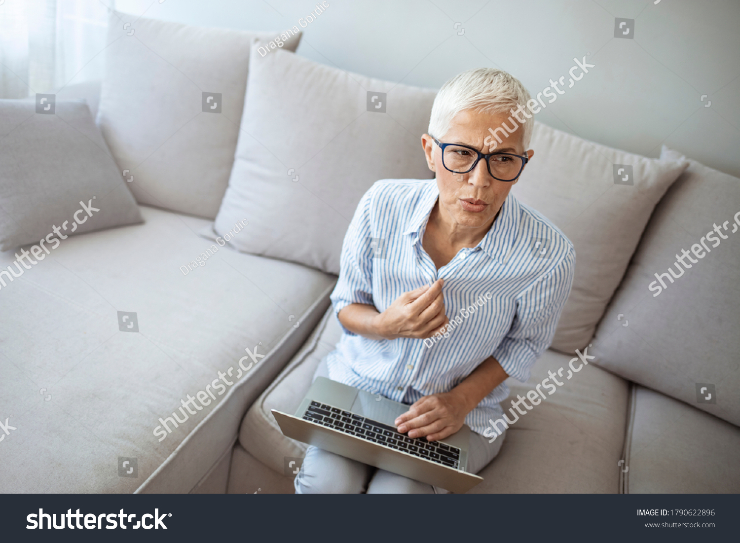 Gradual process.Beautiful mature woman touching shirt and having hot flash. Exhausted mature woman entering menopause. Portrait of an attractive senior woman sitting on a sofa at home with a hot flash #1790622896