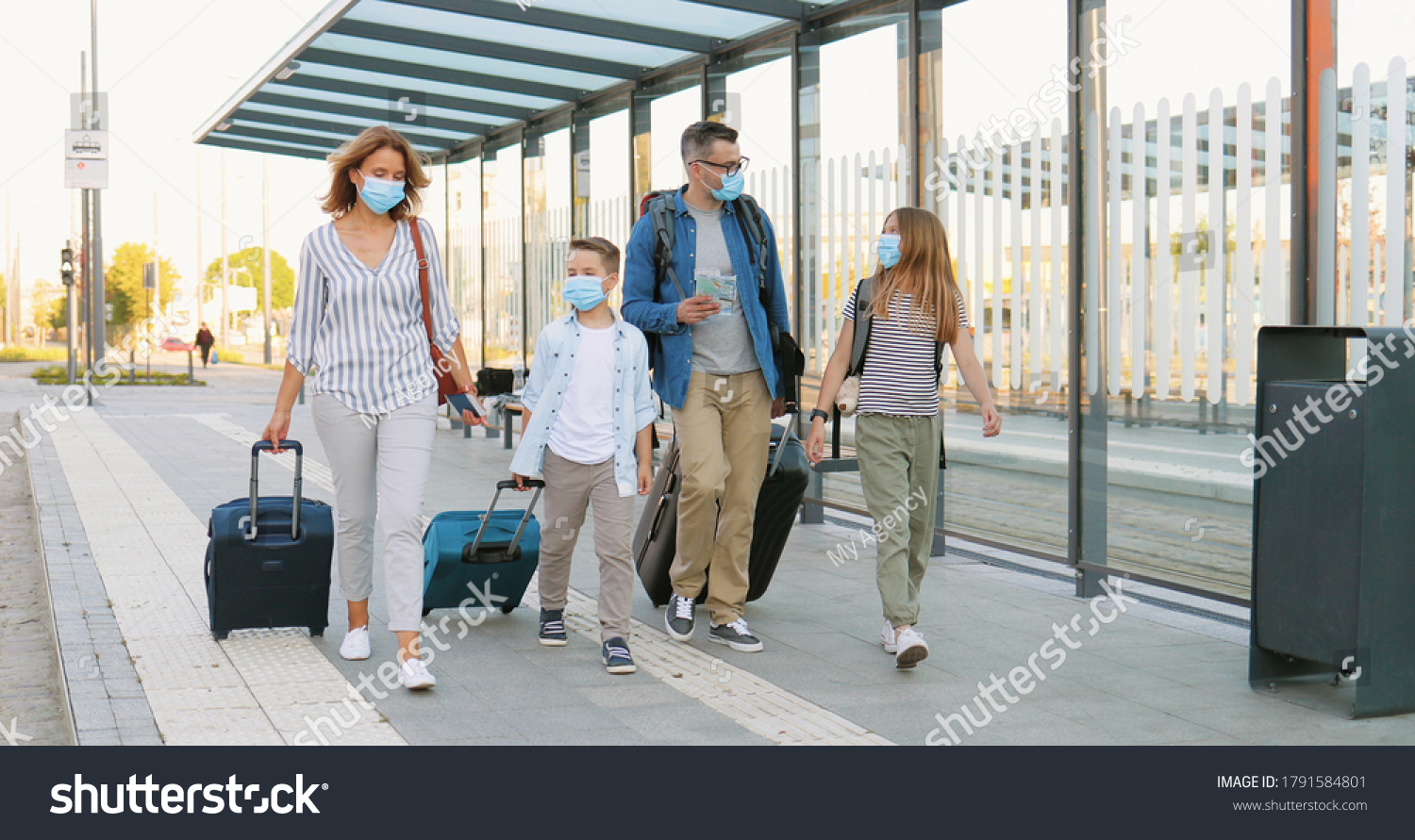 Caucasian happy family with two cute small kids walking at bus stop or train station, carrying suitcases on wheels and talking. Parents with little daughter and son in medical masks travelling. #1791584801