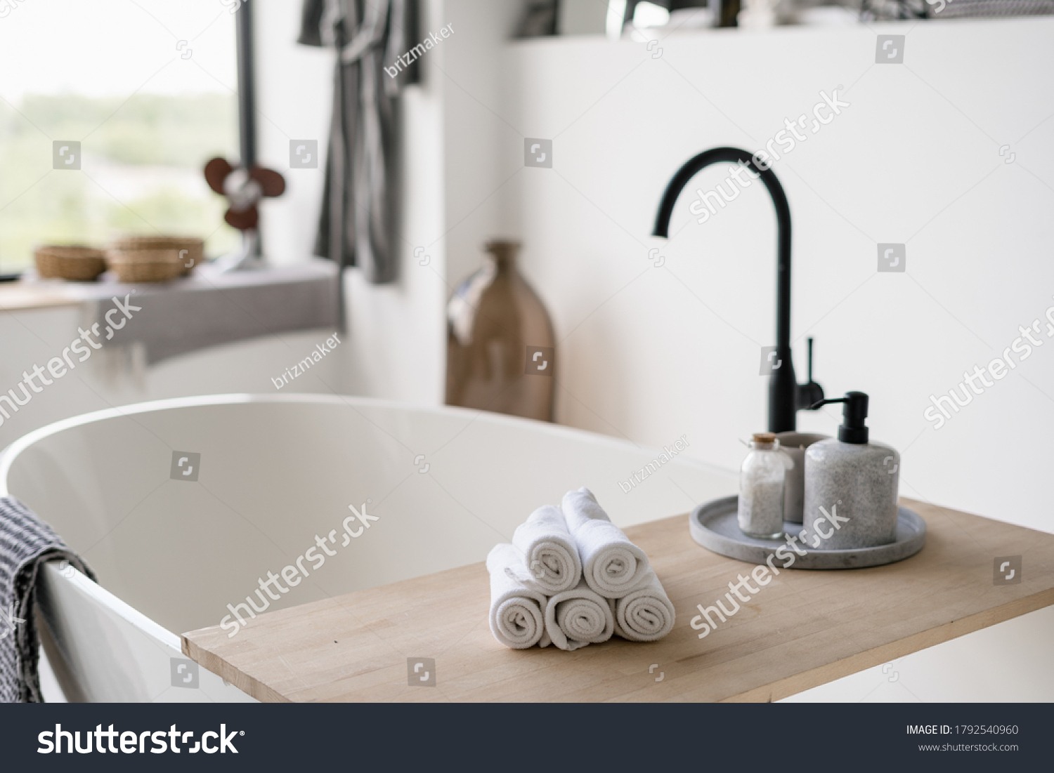 Selective focus on clean folded towels at wooden shelf on empty and contemporary tub with modern black water tap on blurred background. House decor at bright home with white interior design #1792540960
