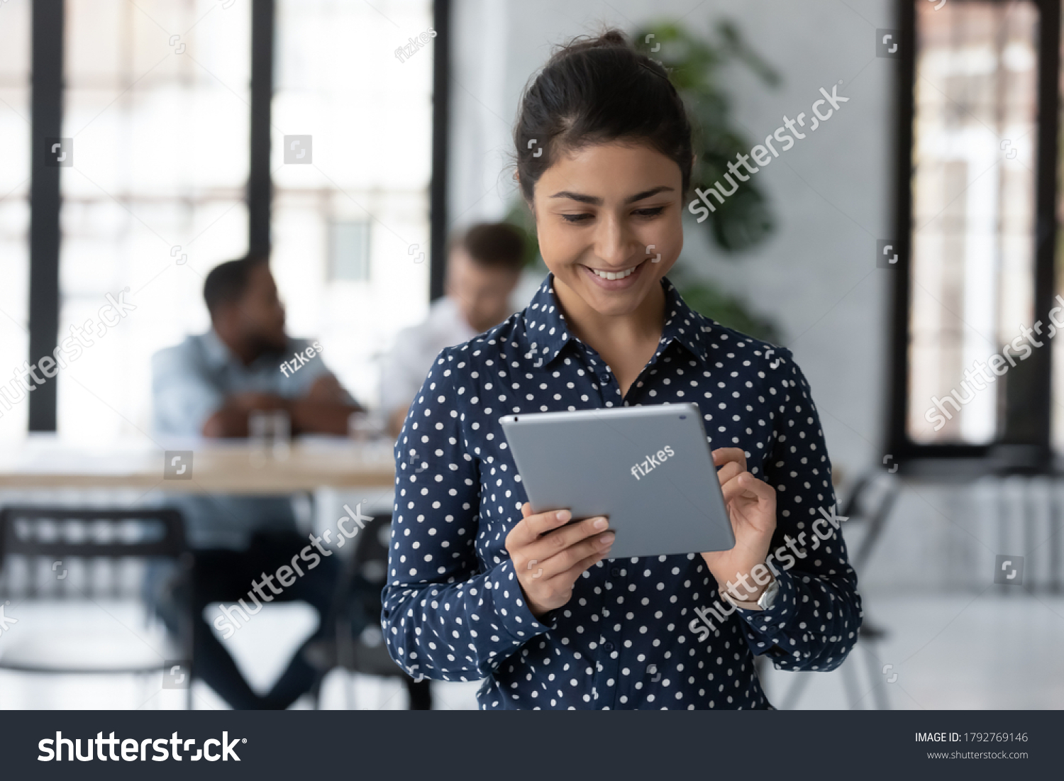 Close up smiling Indian businesswoman holding digital tablet, standing in modern office room, employee working online, using apps, writing or reading email, chatting in social network during break #1792769146