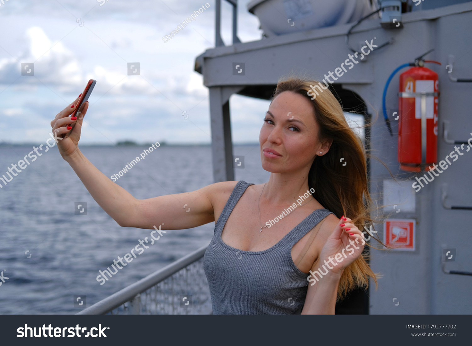 Beautiful white woman with blonde hair in grey dress take a selfie shot and looking to camera when sailing on ferryboat in cloudy day. Small depth of field on background. Traveling on the river. #1792777702