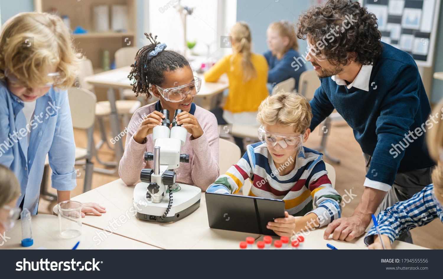 Elementary School Science Classroom: Cute Little Girl Looks Under Microscope, Boy Uses Digital Tablet Computer to Check Information on the Internet. Teacher Observes from Behind #1794555556
