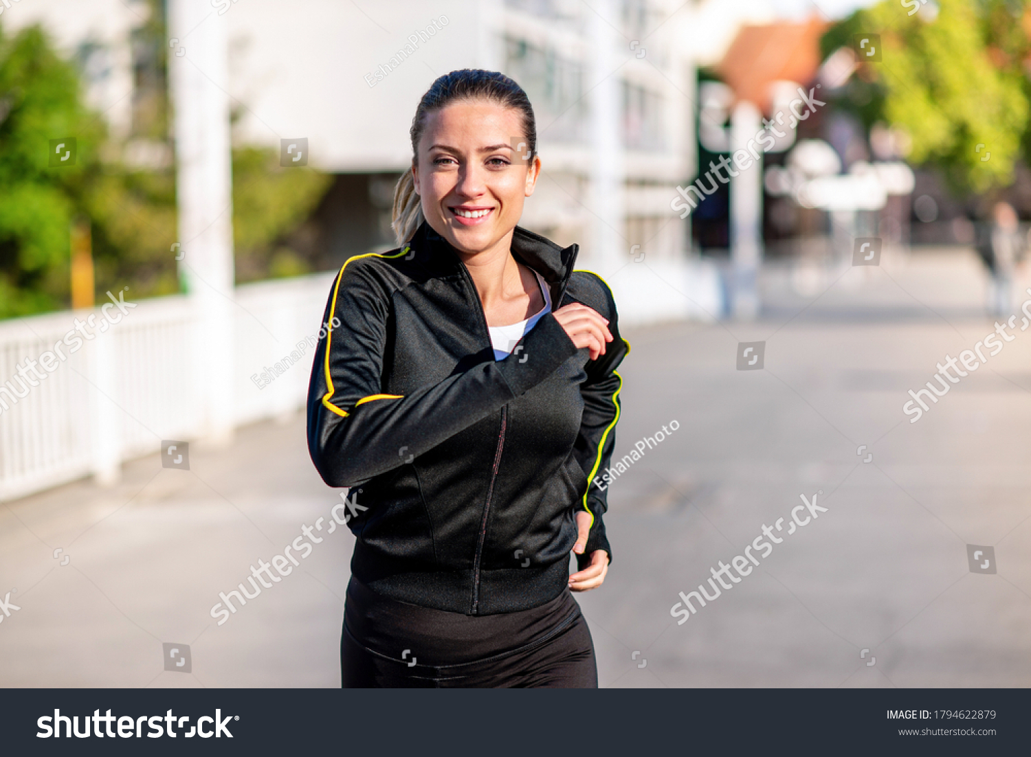 Closeup of a young smiling woman runner in black long sleeve track suit, running across the bridge on a sunny day #1794622879
