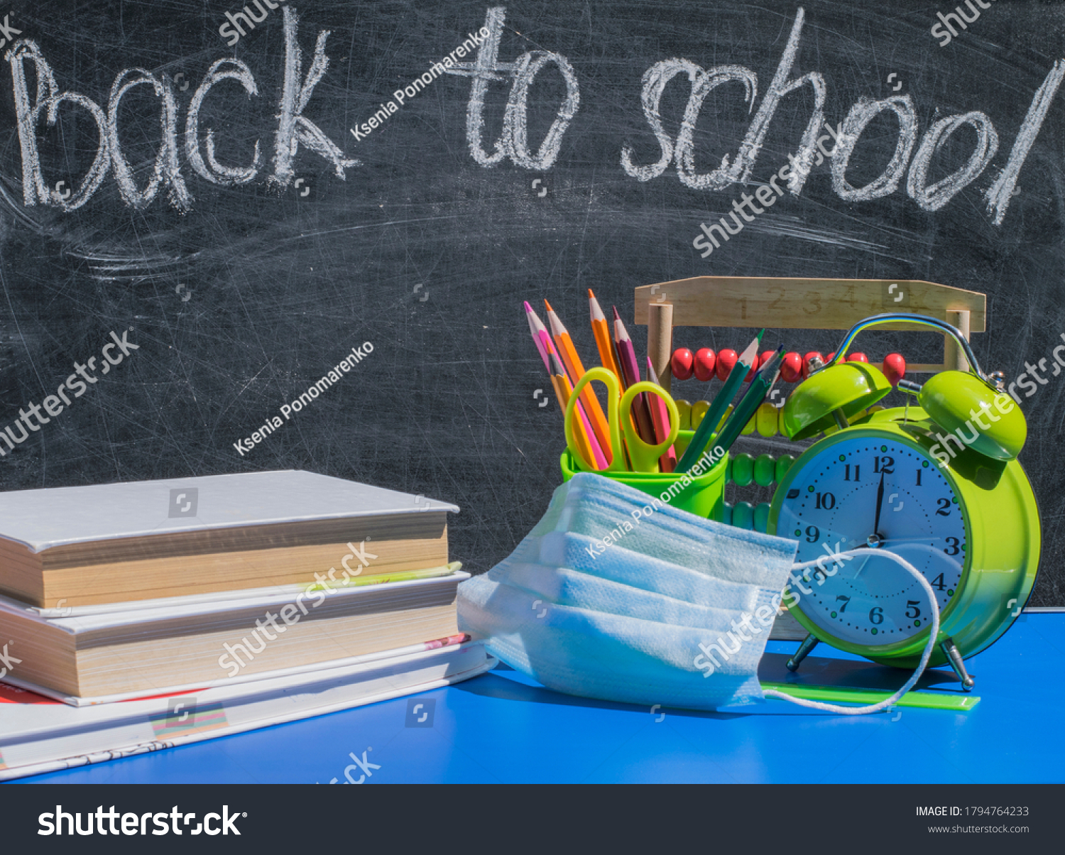 School supplies, alarm clock, books and  medical mask on the school desk against the background of the blackboard with the words "back to school" Concept for a new learning environment in the COVID-19 #1794764233