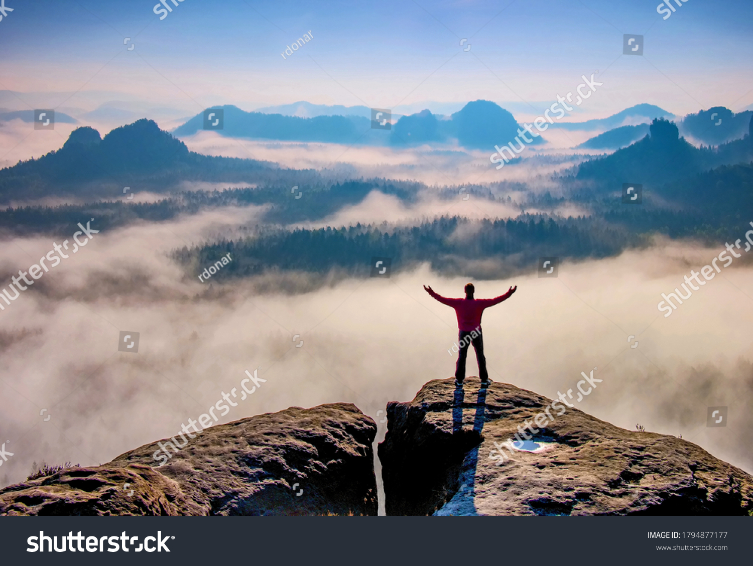 Small silhouette of hiker man enjoying beautiful sunrise in morning mountains. Traveler with raised hands standing on mountain with white fog below. #1794877177