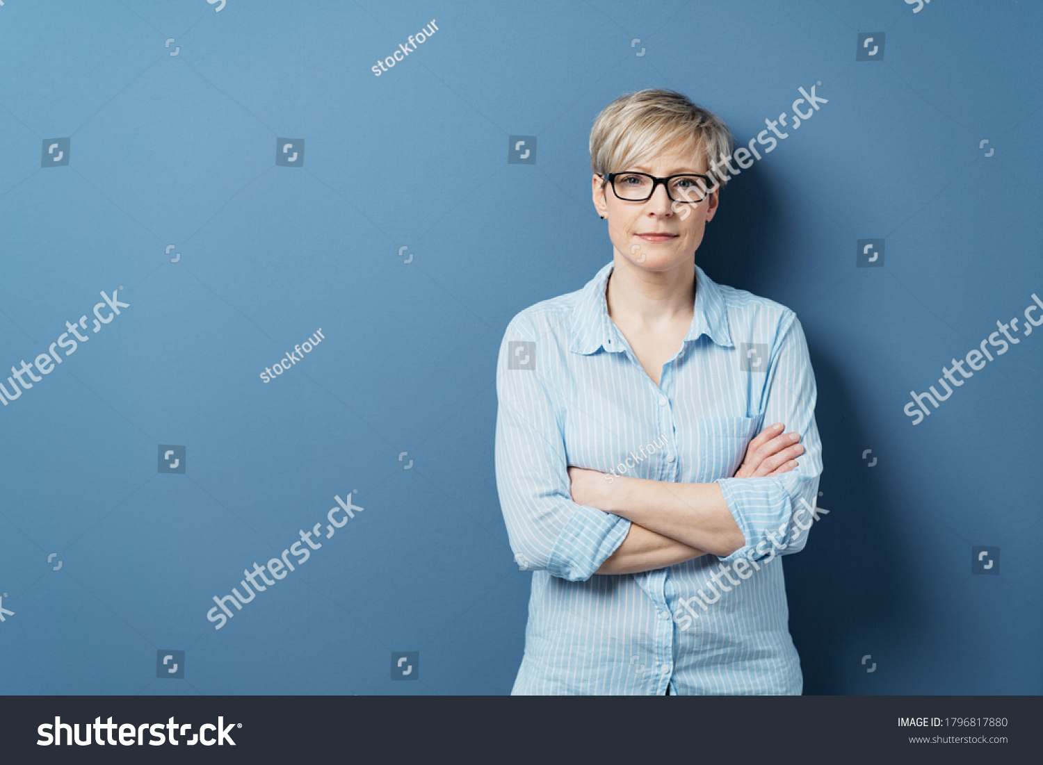 Portrait of mature blonde woman wearing glasses standing against blue background with arms crossed #1796817880