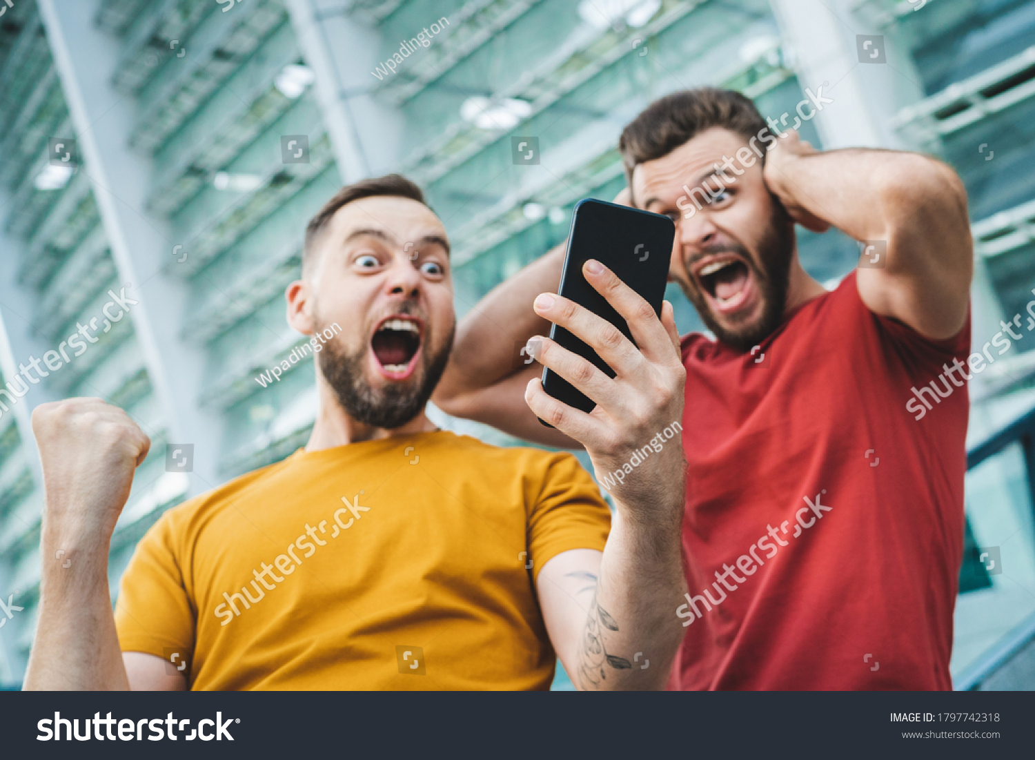 Two friends showing sincere emotions of joy about victory in online lottery. Men being happy winning a bet in online sport gambling application with football stadium on the background. #1797742318