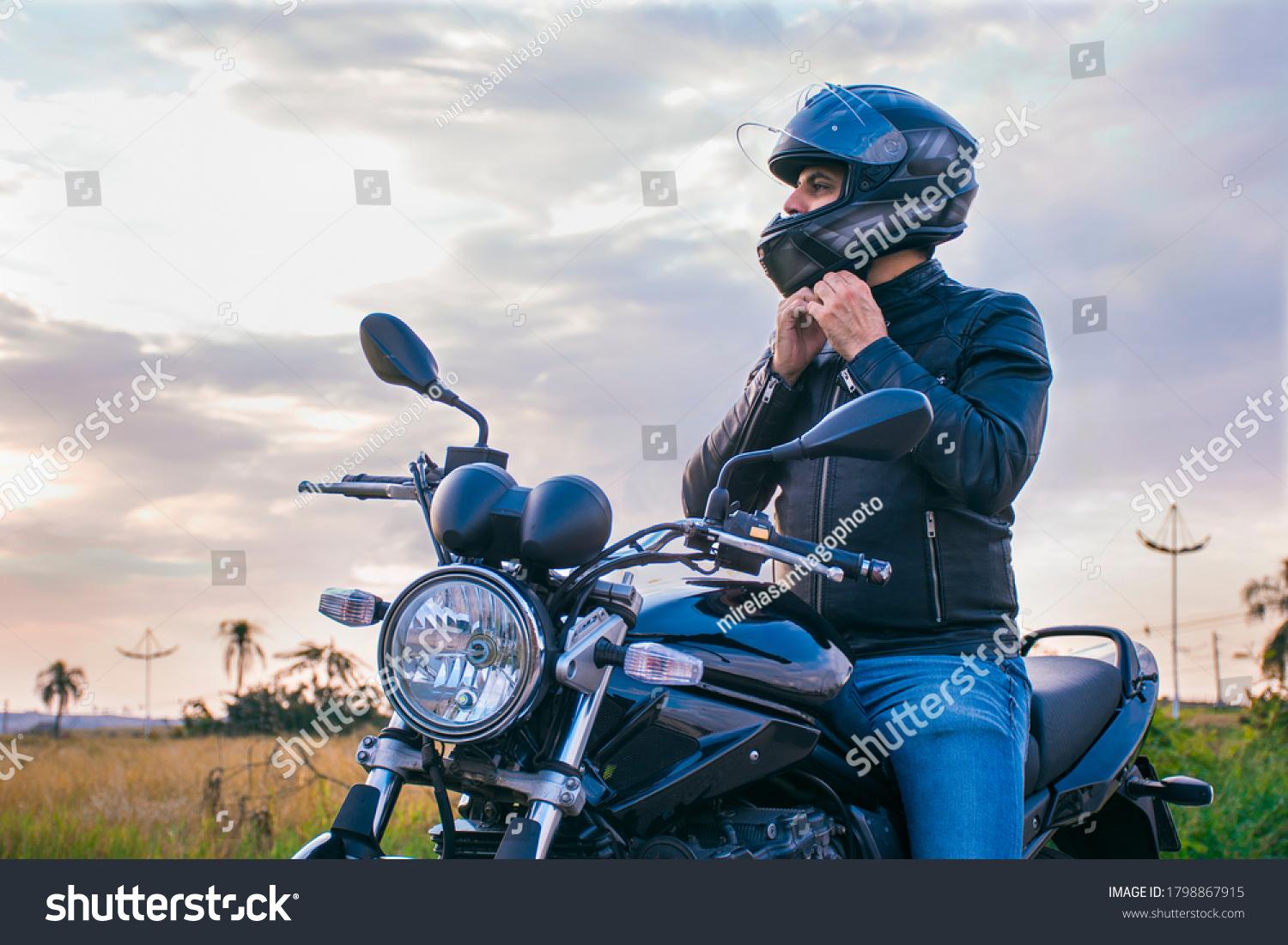 Man sitting on a motorcycle, wearing jeans and a black jacket, fastening his helmet with a landscape in the background. #1798867915