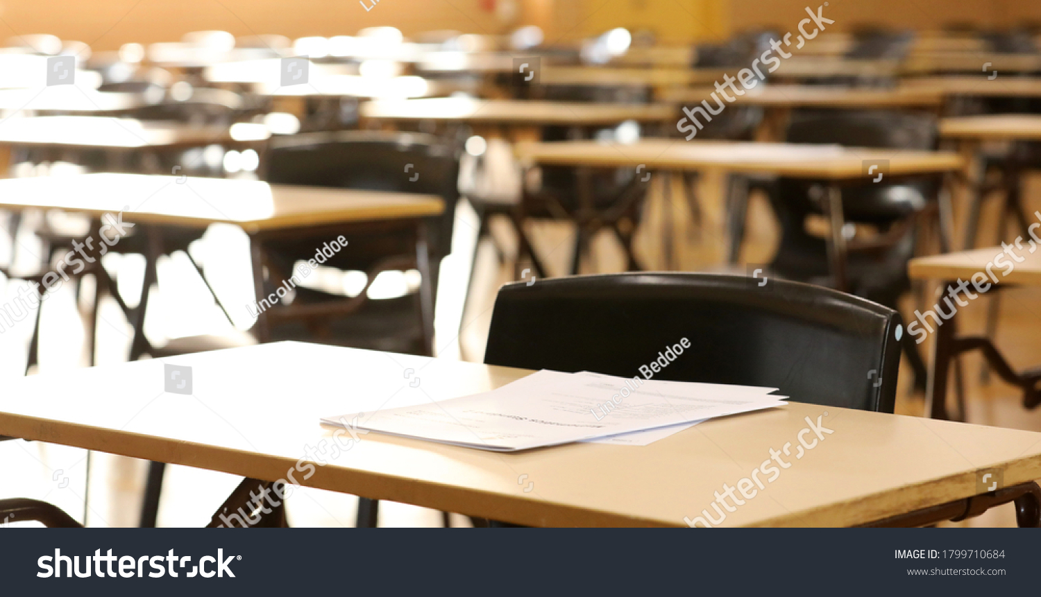 A high school hall or room set up ready for an end of year final exam to be sat by students. examination paper sitting on the edge of a desk or table.  #1799710684