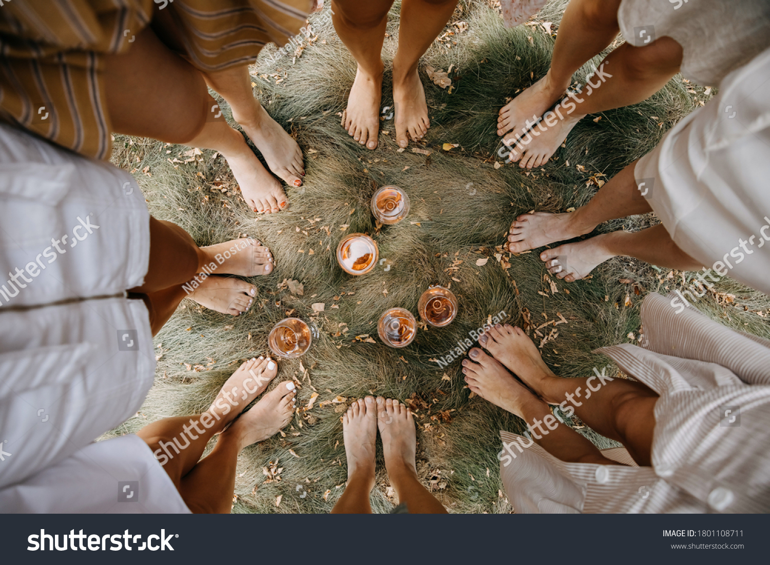 Female barefoot feet on dry grass standing in a circle with glasses of wine in the middle. #1801108711
